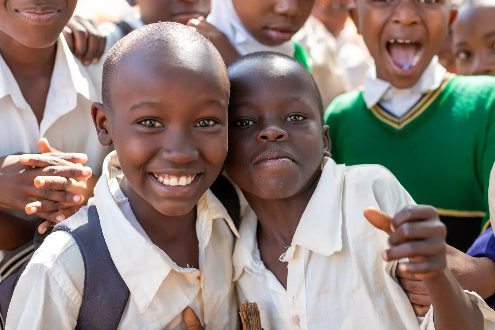 Two grinning students in white school shirts, classmates crowding in behind them